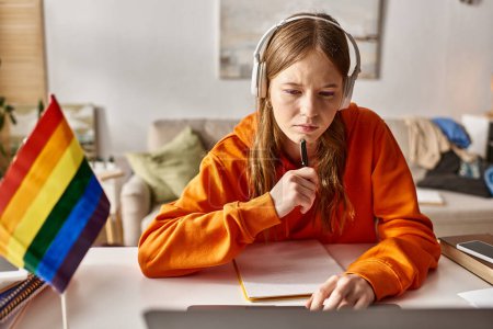 Téléchargez les photos : Concentré jeune adolescente dans les écouteurs sans fil engagés dans l'apprentissage en ligne à côté drapeau de fierté et ordinateur portable - en image libre de droit