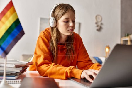 Foto de Joven adolescente enfocada en auriculares inalámbricos escribiendo en su computadora portátil junto a la bandera de orgullo - Imagen libre de derechos