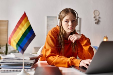 Foto de Cansada joven adolescente en auriculares inalámbricos utilizando su computadora portátil junto a la bandera de orgullo y papelería - Imagen libre de derechos