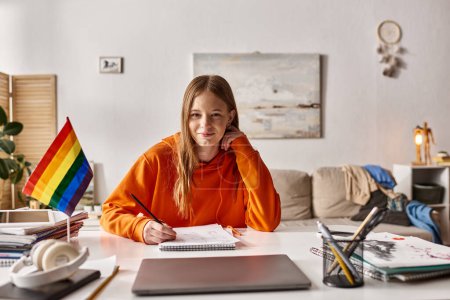 Photo for Cheerful teenage girl drawing a sketch, immersed in creative process with pride flag beside her - Royalty Free Image