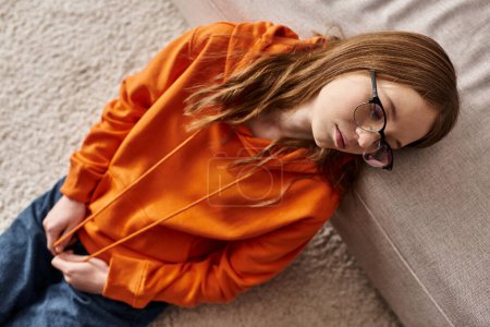 Foto de Vista superior de chica en sudadera con capucha naranja salones cerca de sofá en casa, melancolía y soledad de adolescente - Imagen libre de derechos