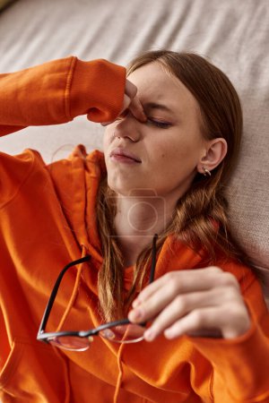 vista superior de la chica adolescente cansada con capucha naranja apoyada en el sofá en casa, melancolía y soledad
