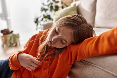 aufgebrachte Teenager-Mädchen in orangefarbenem Kapuzenpullover, die sich auf der Couch in einer gemütlichen häuslichen Umgebung lehnen, Einsamkeit und Traurigkeit