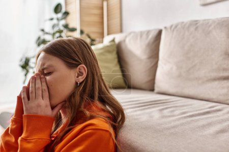 Teenage girl in distress, covering face with hands while crying near couch in living room