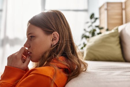 Photo for Side view of teenage girl in distress looking away while sitting near couch in living room - Royalty Free Image