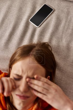 top view of teenage girl in orange hoodie lying near smartphone on couch, stress from social media
