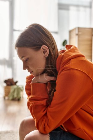 upset teenager girl in orange hoodie sitting on couch in Moden apartment, solitude and sadness