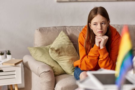 Pensive teen girl in hoodie sits on couch with a distant look, blurred lgbtq flag on foreground