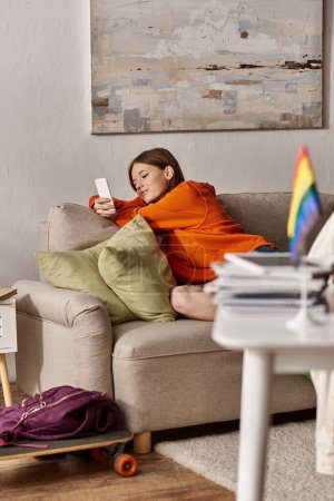 Téléchargez les photos : Joyeux adolescent fille à l'aide de son smartphone près du canapé et flou drapeau lgbt au premier plan - en image libre de droit