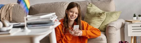 Cheerful teen girl using her smartphone near couch and blurred lgbt flag on foreground, banner