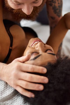 close up of happy multicultural couple laughing together on bed in light-filled bedroom, connection