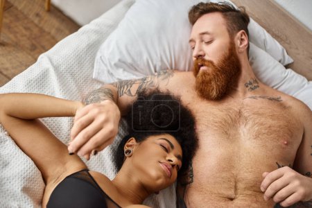 vista superior del hombre barbudo y tatuado acostado en la cama con la novia afroamericana, vinculación