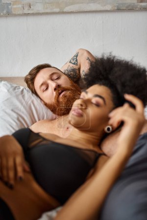 barbudo hombre con tatuaje mirando a su novia afroamericana en lencería acostada a su lado