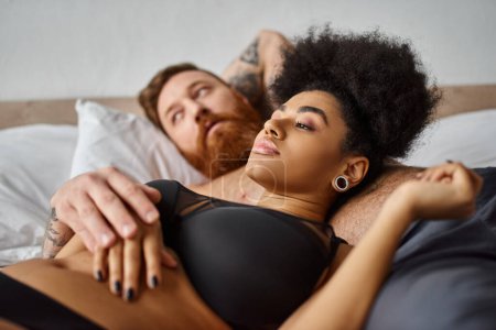 young and curly african american woman in lingerie lying next to her man in bedroom, soulmates