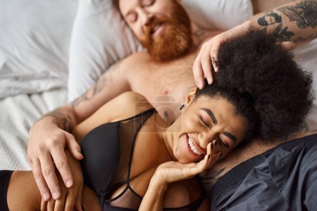 Téléchargez les photos : Souriante et frisée afro-américaine en lingerie couchée à côté de son homme dans la chambre, âmes sœurs - en image libre de droit