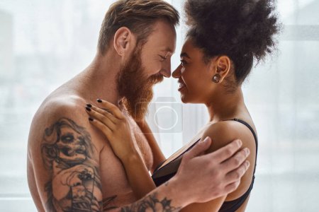 Foto de Hombre barbudo y tatuado feliz sin camisa abrazando a su novia afroamericana cerca de la cortina - Imagen libre de derechos