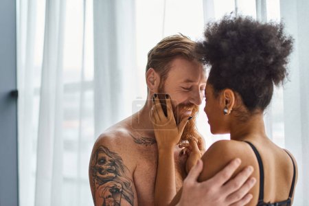 hombre barbudo y tatuado feliz sin camisa abrazando a su novia afroamericana cerca de la cortina