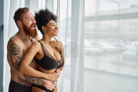 Foto de Barbudo y hombre tatuado sin camisa abrazando a su novia afroamericana feliz cerca de la cortina - Imagen libre de derechos