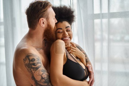 barbudo y hombre tatuado sin camisa besando a su novia afroamericana feliz cerca de la cortina