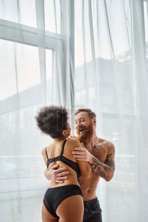excited and tattooed man without shirt hugging his happy african american girlfriend near curtain