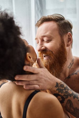excited and tattooed man without shirt hugging curly african american girlfriend near curtain