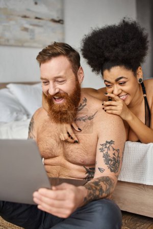 Téléchargez les photos : Heureux tatoué homme et afro-américaine femme regarder film comédie sur ordinateur portable, couple multiculturel - en image libre de droit