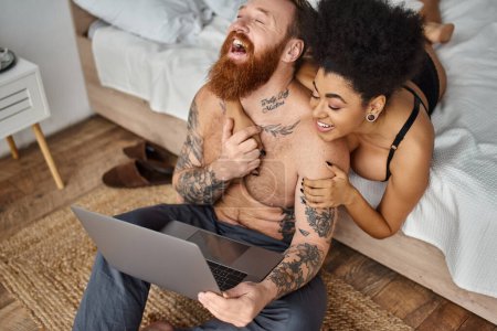 Téléchargez les photos : Homme tatoué et femme afro-américaine regardant un film de comédie sur ordinateur portable et riant ensemble - en image libre de droit