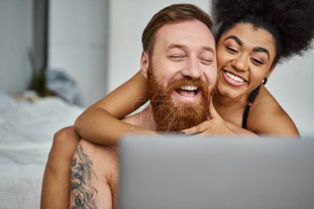 tattooed man and african american woman watching comedy movie on laptop and laughing together