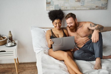 happy african american woman in lingerie using laptop and lying on bed with tattooed boyfriend