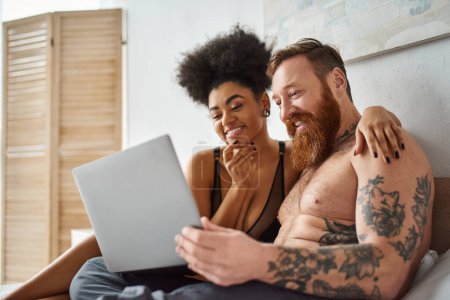 positive african american woman in lingerie using laptop and lying on bed near tattooed man, comedy