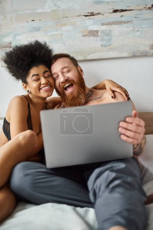 happy african american woman in lingerie using laptop and lying on bed near tattooed man, movie