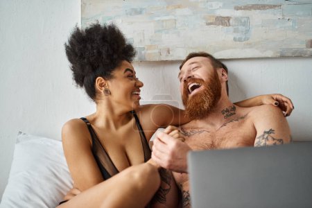 Téléchargez les photos : Heureuse femme afro-américaine en lingerie à l'aide d'un ordinateur portable et couché sur le lit près d'un homme tatoué, rire - en image libre de droit