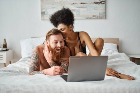 Téléchargez les photos : Interracial couple regarder la comédie film sur lit, afro-américaine femme et barbu tatoué homme - en image libre de droit