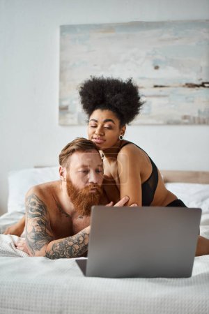interracial couple watching movie on bed, curly african american woman and bearded tattooed man
