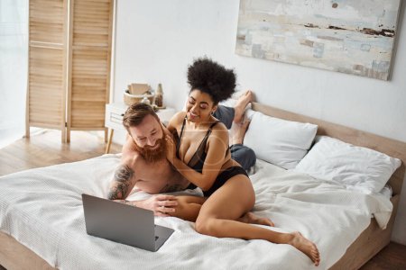 Téléchargez les photos : Interracial couple regarder film sur lit, frisé afro-américain femme et barbu tatoué homme - en image libre de droit