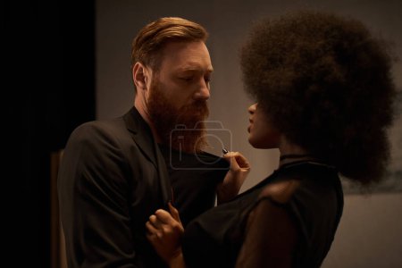 curly african american woman in dress pulling blazer of bearded man during date, sexy couple