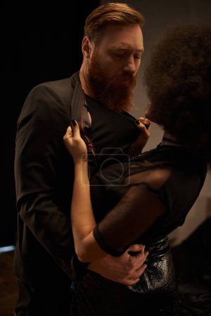 young african american woman in dress pulling blazer of bearded man during date, sexy couple
