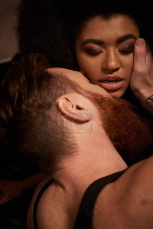 Téléchargez les photos : Homme barbu avec des tatouages touchant le visage de la femme afro-américaine bouclée tout en la séduisant sur le lit - en image libre de droit
