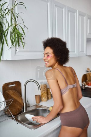 mujer afroamericana feliz y rizada en lencería con parches rosados bajo los ojos lavando platos