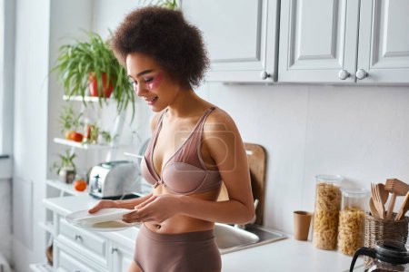 happy and curly african american woman in lingerie with pink patches under eyes holding clean dishes