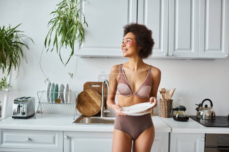 happy and curly african american woman in lingerie with pink patches under eyes holding clean dishes
