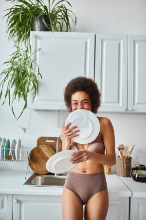 happy african american girl in lingerie with pink patches under eyes covering face with clean dishes