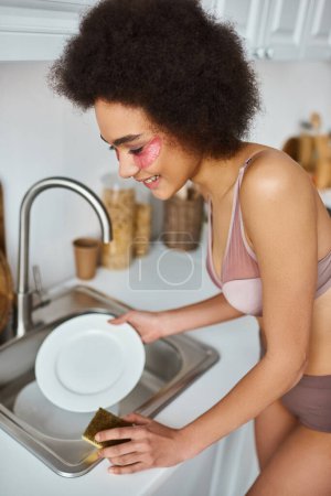 mujer afroamericana en sujetador con parches rosados bajo los ojos sonriendo y lavando plato con esponja