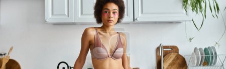 curly african american woman in bra with pink patches under eyes standing in modern kitchen, banner
