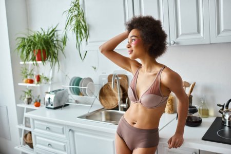 smiling african american woman in bra with pink patches under eyes standing in modern kitchen