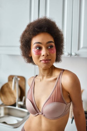 portrait of african american woman in bra with pink patches under eyes standing in modern kitchen