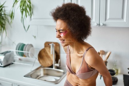 portrait of happy african american woman in bra with pink patches under eyes laughing in kitchen