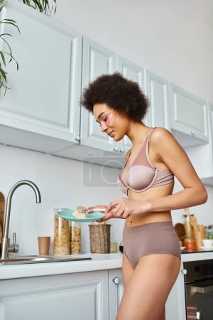 pretty african american woman in bra with pink patches under eyes washing plate with dish brush