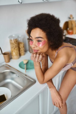 mujer afroamericana complacida en sujetador con parches rosados bajo los ojos sonriendo en la cocina