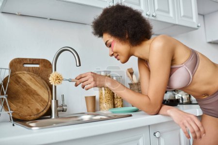 mujer afroamericana complacida en sujetador con parches rosados bajo los ojos sosteniendo la esponja plato en la cocina